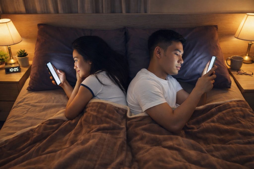 A couple lying in bed using their phones instead of interacting, showing digital disconnection in relationships.