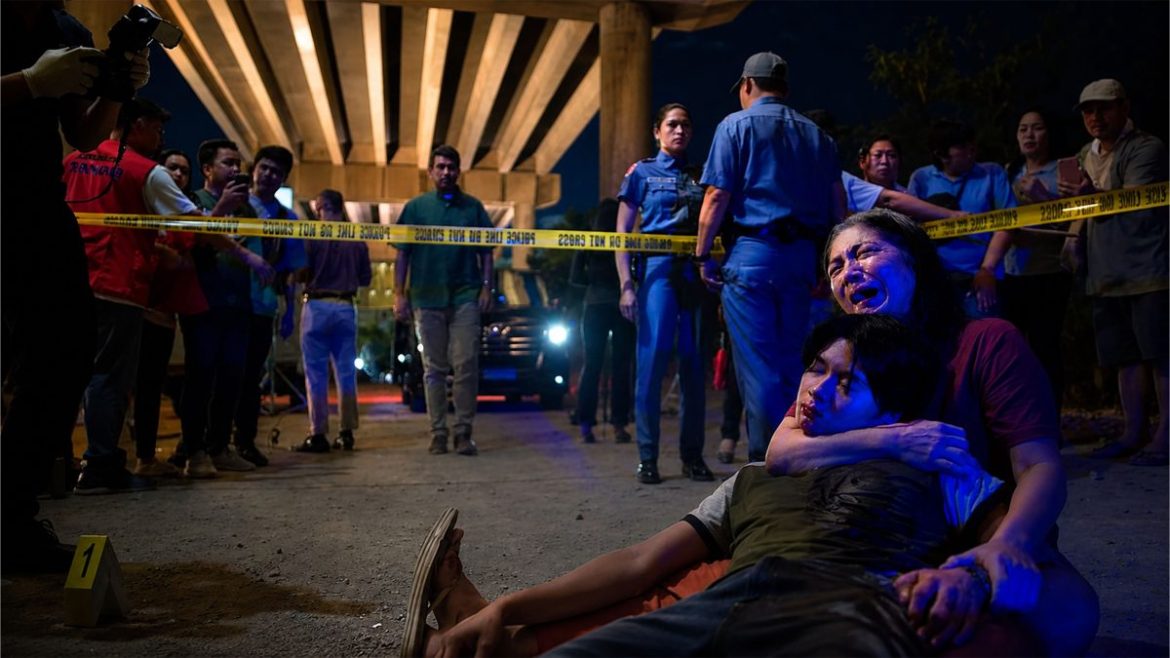 A grieving woman holds a young man at a crime scene surrounded by police officers and bystanders in the Drug War trailer.