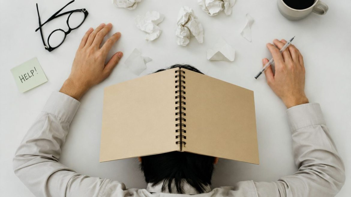 Overhead view of an exhausted office worker covering their face with a notebook while sitting at a cluttered desk.