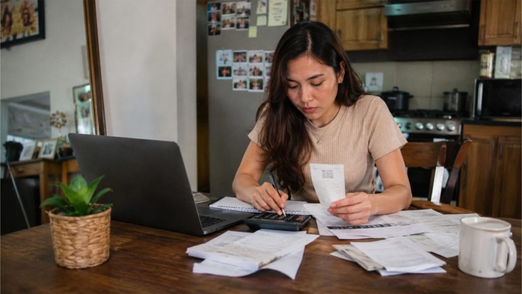 Filipino woman calculating expenses and checking receipts beside a laptop