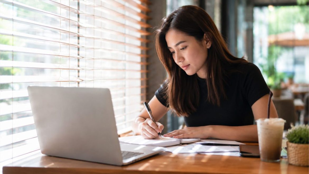 Filipino woman working on a laptop while planning a side hustle business and reviewing notes at a café.