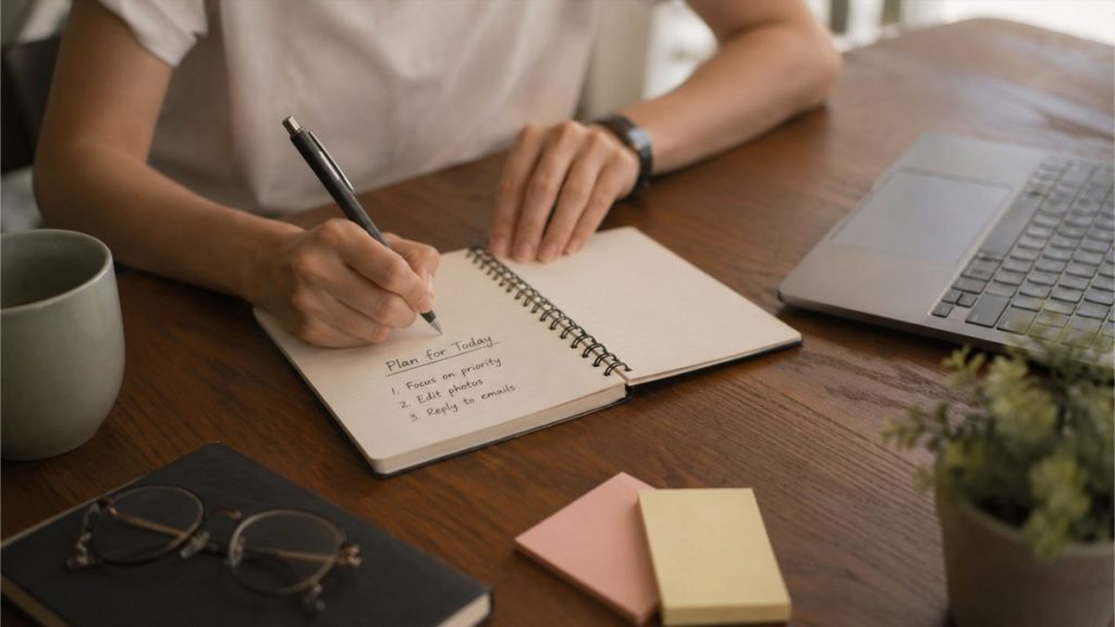 Individual organizing daily tasks in a notebook beside a laptop and work desk